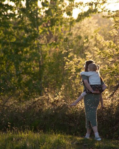 Natürlicher Moment: Mutter geht im Wald mit Tochter auf dem Rücken, Sonnenlicht fällt durch die Bäume – Familienfotoshooting Kiel