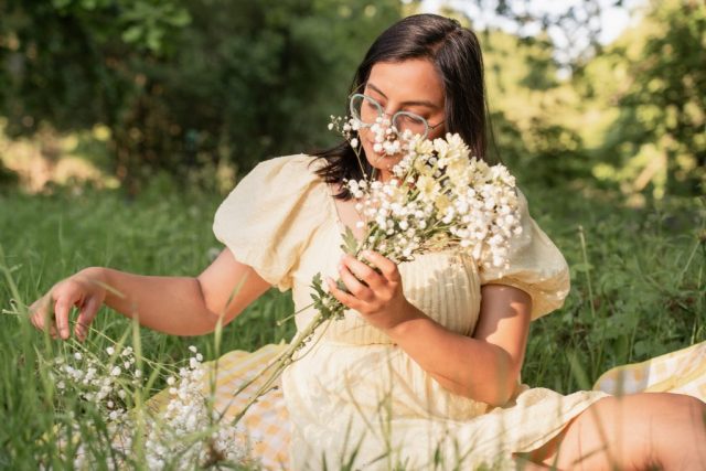 Frau hält zarte Blumen in der Hand und sitzt entspannt im Gras unter Bäumen – Portraitfotoshooting bei Abendlicht in Kiel