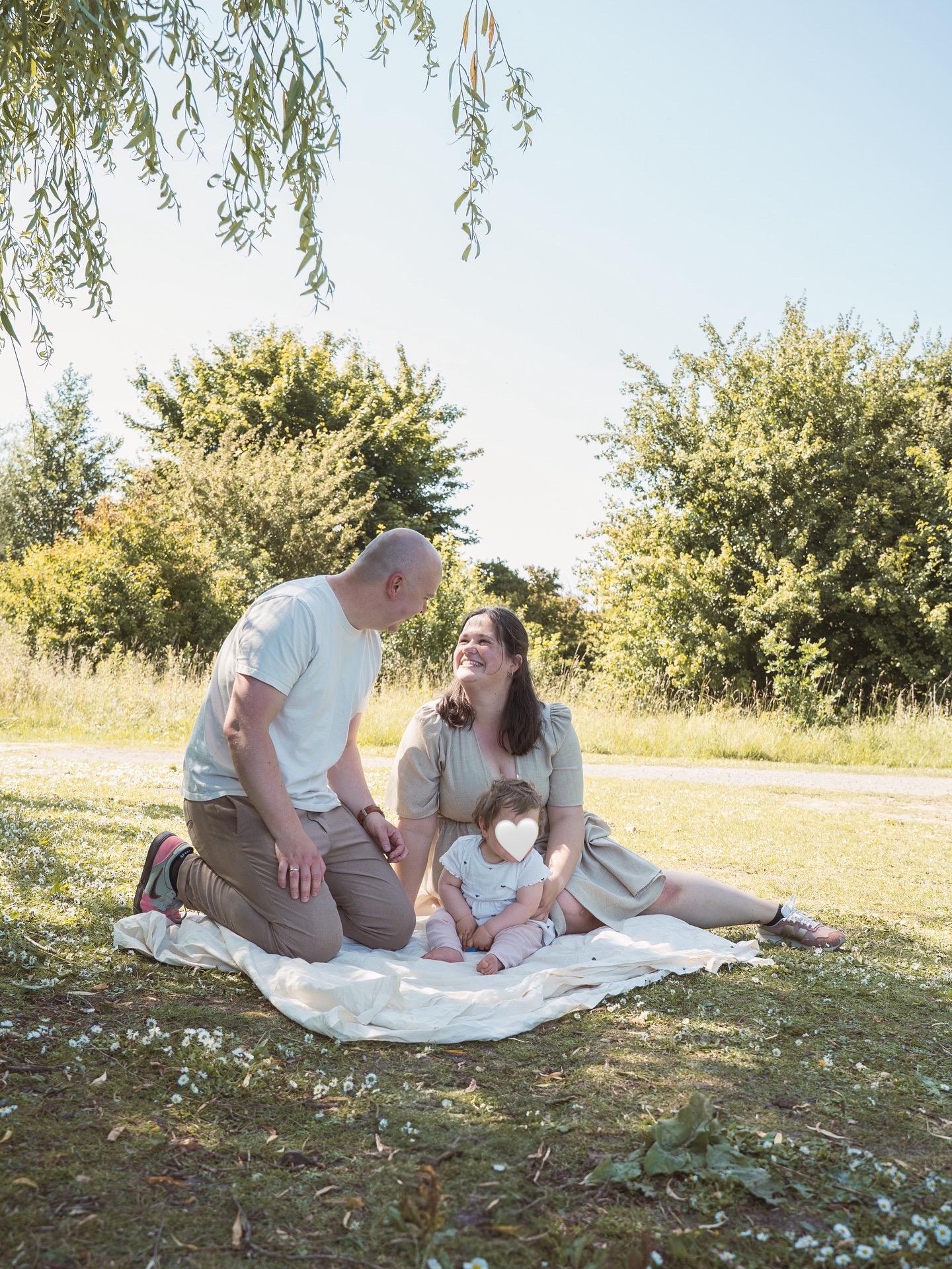 Familienfoto im Park in Kiel: Vater, Mutter und ihre kleine Tochter sitzen gemeinsam auf einer Leinen-Decke im Gras und genießen die Zeit miteinander