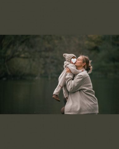 Liebevoller Moment beim Familienfotoshooting in Kiel: Mutter hebt ihre kleine Tochter in die Luft und küsst sie auf die Wange am See im Park