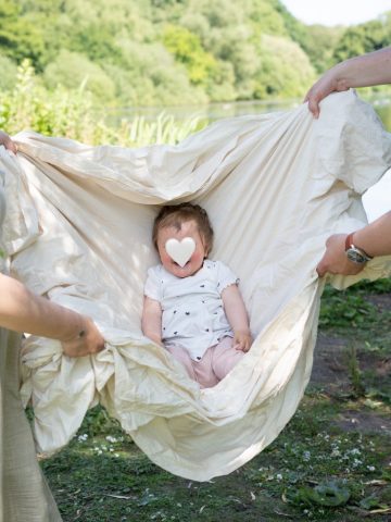Kleines Mädchen wird von Mutter und Vater im Leinentuch geschaukelt und freut sich – lebendige Familienaufnahme aus Kiel