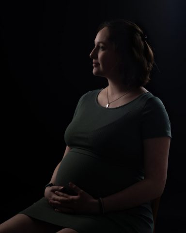 Schwangere Frau sitzt auf einem Stuhl im Studio und blickt ruhig zur Seite, das kunstvolle Licht betont ihren Bauch